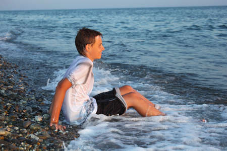 sitting teenager boy in wet clothes on stone seacoastの写真素材