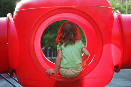 little girl sits in red plastic box on playground, view from backの写真素材