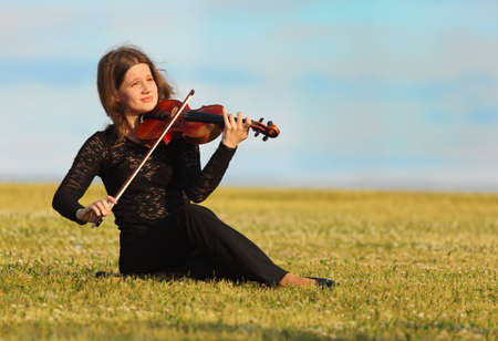 girl sits on grass and plays violin against  skyの写真素材