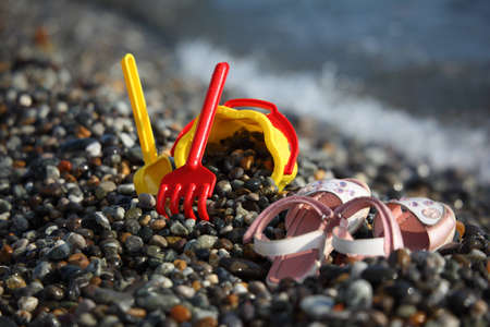 Yellow children's bucket with a scoop, a red rake and sandals on seacoastの写真素材
