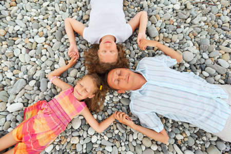 Happy family with little girl lying on stony beach, closed eyes, having joined hands, Concerning with headsの写真素材
