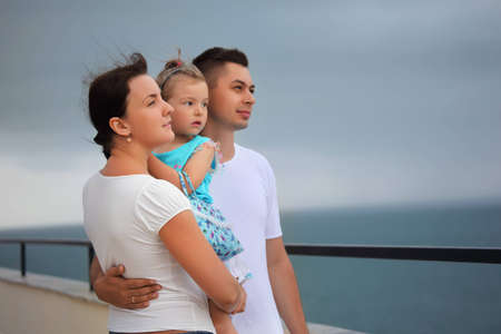 young man and beautiful woman protects little girl from wind on seacoastの写真素材