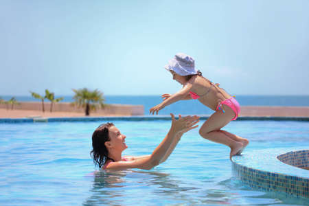 beautiful woman catches little girl jumping in pool against seaの写真素材