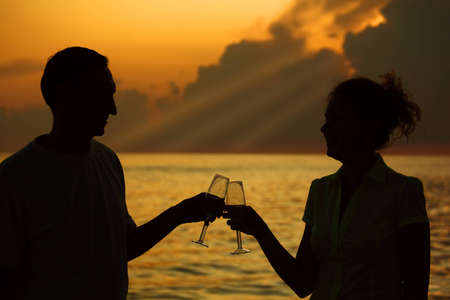 Man and woman clink glasses. Silhouettes against sea. Rays of lights on sky.の写真素材