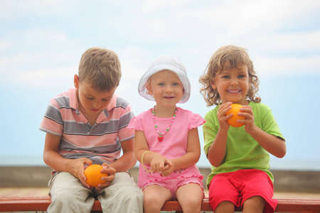 boys and girl with oranges and one liittle girl with panama hat is sitting on wooden bench. boy wearing stripped t-shirt looking at orange. focus on face of girl with orange.の写真素材