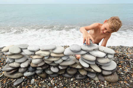 little boy is building construction of many pebbles on a sea coast near water.の写真素材