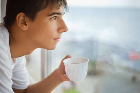 teenager boy with cup in hand looking out of window in morning, focus on cup
の写真素材