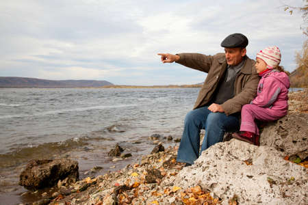 Grandfather and granddaughter sit on stone and look at lakeの写真素材