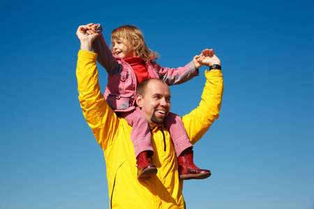 Cheerful little girl sits on shoulders at man, against blue sky.の写真素材