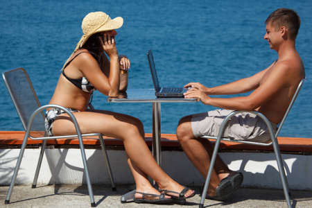 Man and woman sitting at table under open sky on background of sea. Man works for laptop. Girl talking on the phone.の写真素材