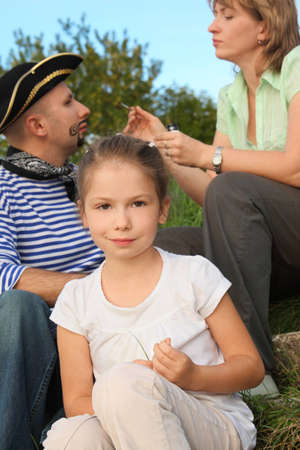 family in early fall park. little girl in focus. woman drawing funny whiskers and beard on man's face in out of focus.の写真素材