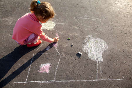 girl sits on concrete asphalt square road. girl draws painting line sun house tree a chalk on asphalt. chld drawings paintings on asphalt concrete. back side viewの写真素材