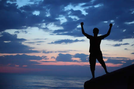 Silhouette guy lifted hands upwards on breakwater in evening near seaの写真素材