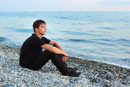 sitting teenager boy on stone seacoast, Looking afarの写真素材