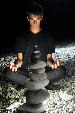 teenager boy meditating near pyramid from pebble on stony seacoast at nightの写真素材