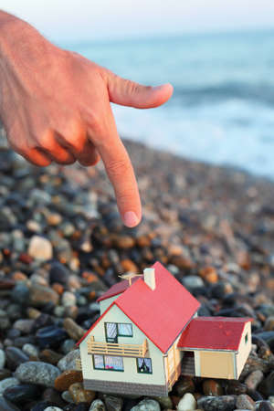 model of house with garage on stony beach in evening, Man's hand with forefinger over houseの写真素材