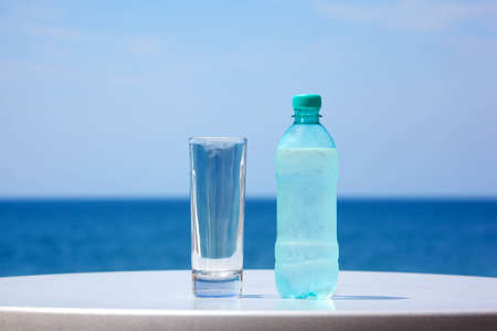 Bottle of water and glass on table under open sky on background of sea.の写真素材