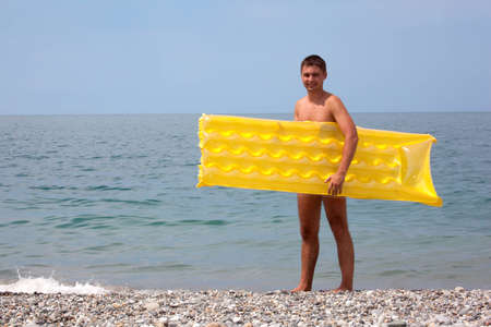 young guy with yellow mattress on seacoast. Man stands on pebble beachの写真素材