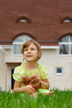 little girl sitting on lawn in front of new home. In her hands she holds toy wolf. vertical formatの写真素材