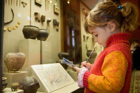 little girl writes to writing-books at excursion in historical museum near exhibits of ancient relics in glass cases little girl writes to writing-books at excursion in historical museum near exhibits of ancient relics in glass cases