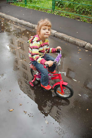 little girl goes on a bicycle on wet asphalt, reflexion in puddleの写真素材