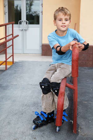 Boy in rollerskates, knee and elbow pads, sitting on the railing in front of house.