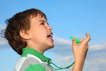 The small beautiful boy, against the blue sky, plays with a green whistle. Profile side.