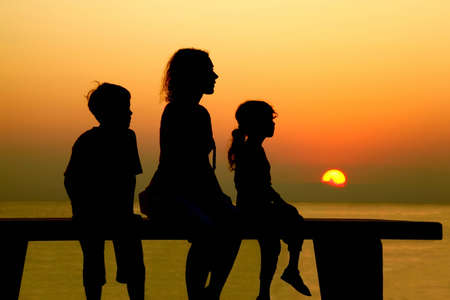 Mother with two children sits on  bench and looks at  beach toward sunsetの写真素材