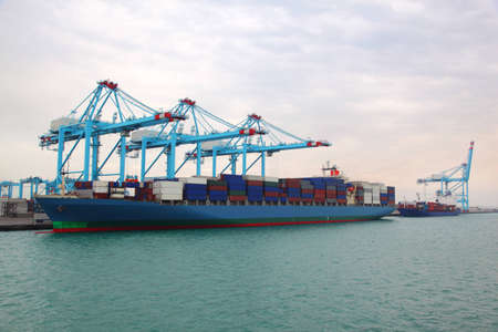Big cargo boat and small barge docked to industrial port with blue cranes