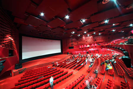High-angle view of screen and rows of comfortable red chairs filled by people in illuminate red room cinemaのeditorial素材