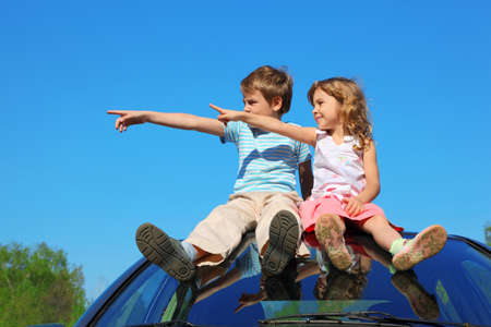 little boy and girl sitting on car roof on blue sky, looking to left side and showing by fingerの写真素材