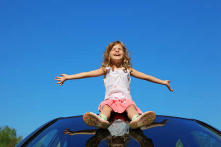 little girl sitting on car roof with open hands on blue skyの写真素材