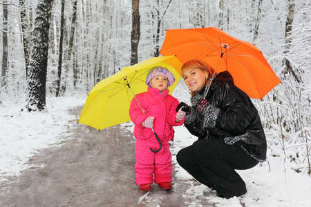 Grandmother and little granddaughter with yellow and orange umbrella stand in woods in snowの写真素材