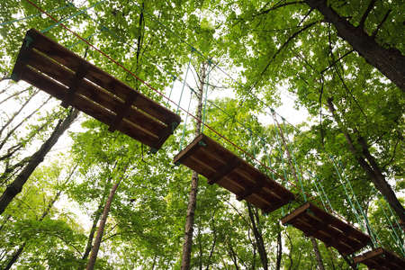 Dangerous ropeway with tether in rope park, trees with green leaves and skyの写真素材