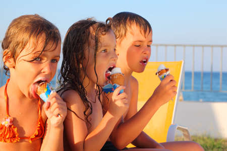 little brother and two sisters in swimsuits on beach eating ice cream after bath. focus on girl in middleの写真素材