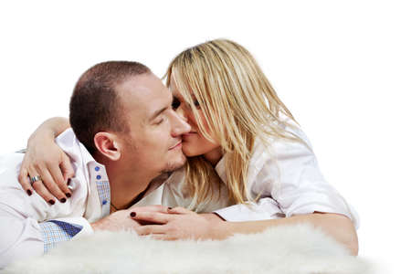 The young pair in white shirts embraces and rubs  their noses, lying on a white fleecy carpet.の写真素材