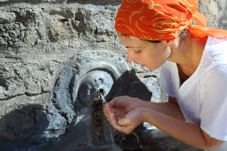beautiful young woman in red kerchief drinking water from small fountain in Vatican wallsの写真素材