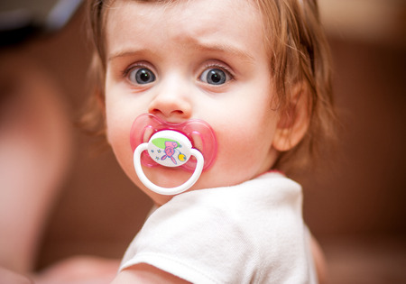 little girl with a pacifier stands near the brown couchの写真素材