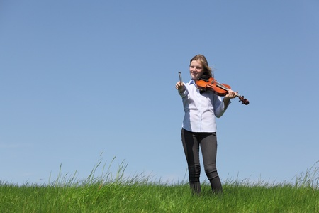 Teenage girl playing violin on meadow over blue sky の写真素材