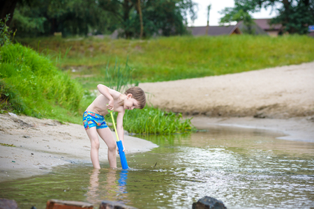 Portrait of Caucasian little boy playing toys and water pump on the river beach. Active leisure for children outdoors.の写真素材