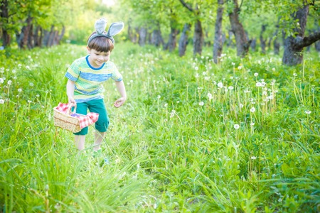 cheerful kid holding basket full of colorful easter eggs standing on the grass in the park after egg hunt.の写真素材