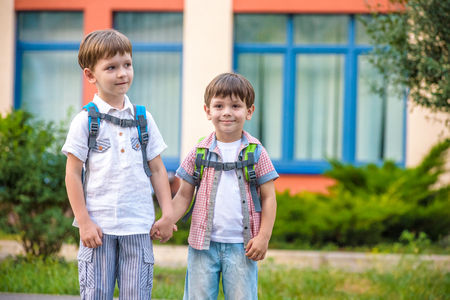 Young students, two sibling brothers, going to school. They hold hands. Children behind shoulders have satchels. Warm day in an early autumn. Back to school. Happy smiling kids.の写真素材