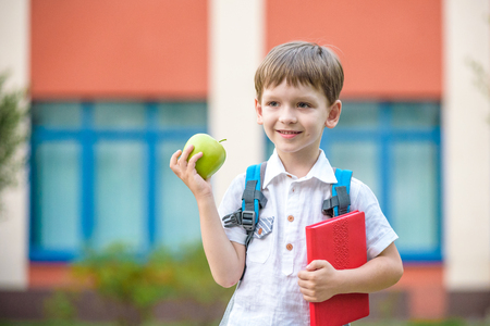 Happy little preschool kid boy with book and green apple. Boy behind shoulders have backpack. First day to school or nursery. Warm day in an early autumn. Back to school concept.の写真素材