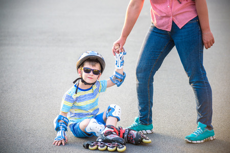 Preschooler falls over while rollerblading with mother in the park. Little boy learning to roller skate. Children wearing protection pads for safe ride. Active outdoor sport for kids.の写真素材