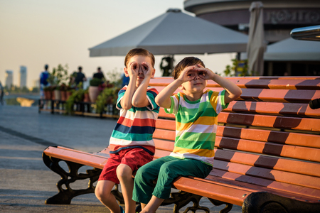 Toddlers boy and his sibling brother sitting on a bench by the city and look for something interesting. Summer time hot evening, hugging each other, friendship concept.の写真素材
