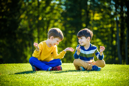 Two Caucasian boy brother friends sitting on fresh green grass field and make meditation dressed in casual clothes, happy kids enjoying peaceful summer weekend outdoors. Friendship relaxation concept.の写真素材