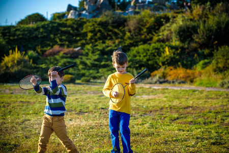 Two active preschool boy playing badminton in outdoor court in summer. Kids play tennis. School sports for children. Racquet and shuttlecock sport for child athlete. Friends happy together warm field.の写真素材