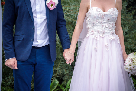 Back view of bride in white dress and groom in suit holding each others hands outdoors.の写真素材
