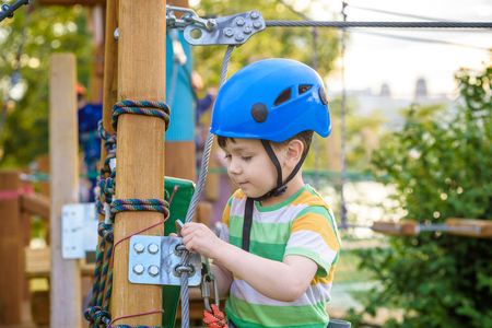 Little cute boy enjoying activity in a climbing adventure park on a summer sunny day. toddler climbing in a rope playground structure. Safe Climbing extreme sport with helmet and Carabiner. insuranceの写真素材