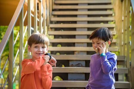 Two friends calling each other while standing on stairs outdoor. One boy has mobile phone, other has smart watch with GPS. Protection and safe communication with children concept.の写真素材